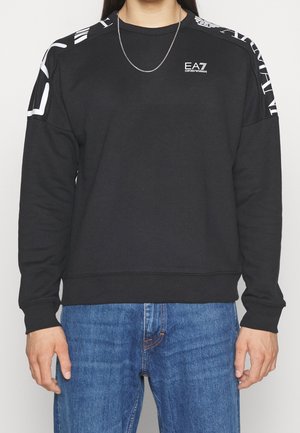 Mężczyzna w czarnej bluzie EA7 Emporio Armani z białym logo na piersi i białymi paskami z logo na ramionach, połączony z niebieskimi dżinsami.