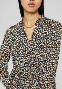 Blouse à imprimé floral noir, mettant en avant de petites fleurs multicolores, un col, et des manches longues avec une texture froncée au niveau du décolleté.
