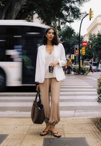 Femme en blazer blanc et pantalon beige se tient à un passage piéton en tenant une tasse de café et un sac fourre-tout alors qu’un bus passe derrière elle.