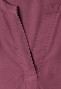 Gros plan d’un vêtement en tissu mauve avec un col en V et des coutures visibles.