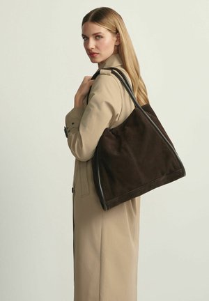 Kobieta o długich blond włosach, ubrana w beżowy trencz, niosąca na ramieniu dużą ciemnobrązową zamszową torbę typu tote.