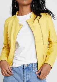 Kvinna iklädd en ljusgul zipjacka över en vit T-shirt och blå denimjeans, accessoriserad med stora guldörhängen.