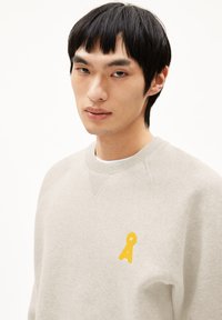 Jonge man met zwart haar in een lichtbeige sweater met een klein geel sleutelvormig logo op de borst, neutrale achtergrond.