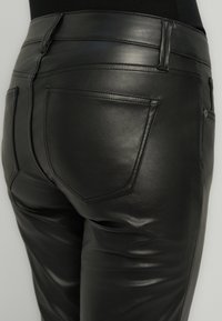 Pantalon en cuir noir à coupe ajustée, présentant une texture lisse, des poches arrière et une taille haute. Les coutures sont visibles le long des coutures.