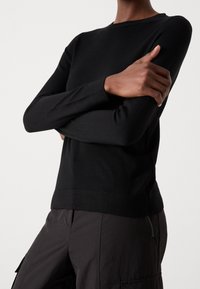 Personne portant un pull noir à manches longues et un pantalon foncé avec une poche zippée, les bras croisés sur la poitrine.