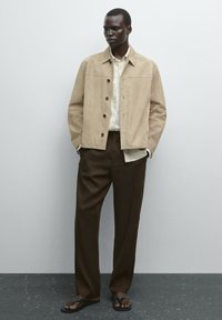 Beige Wildlederjacke mit Kragen und Knopfleiste, kombiniert mit einem hellen karierten Hemd und braunen weiten Hosen. Schwarze Flip-Flops.