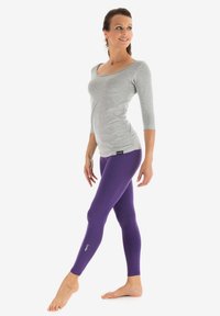 Grå tætsiddende bluse med trekvartærmer parret med lilla leggings, begge lavet af strækbart materiale. Bemærkelsesværdigt logo på leggingsene.