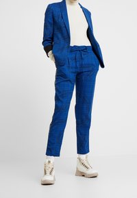 Costume à carreaux bleu avec accents noirs, comprenant une veste cintrée et un pantalon à cordon de serrage, associé à des baskets montantes beige.