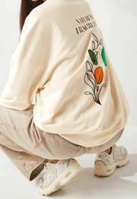 Beżowa, oversizeowa bluza z nadrukiem na plecach przedstawiającym zielone i pomarańczowe ilustracje botaniczne. Połączona z jasnobeżowymi spodniami i trampkami.