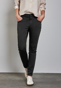 Schwarze Slim-Fit-Hosen mit Taschen vorne, kombiniert mit einem hellbeigen Hemd. Akzentuiert mit einem Knopfverschluss und getragen mit weißen Sneakern.