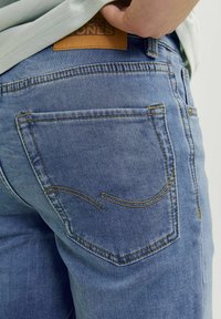 Vaaleansiniset farkut, joissa on sileä pinta, keltainen ompelu ja kaksi takataskua, joissa on kaareva sauma. Nahkainen etiketti vyötäröllä.