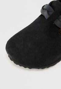 Fekete velúr cipő, fonott fekete pánttal és enyhén texturált talppal, sima fehér háttér előtt fényképezve.