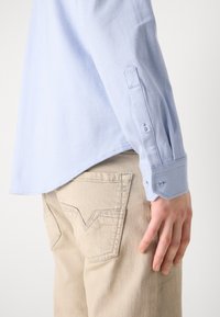Lichtblauw overhemd met knopen, gemaakt van een textuurstof en met een schuine manchette, gecombineerd met beige broek met een zigzagzakdesign.