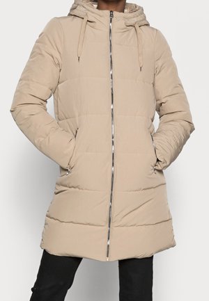 Manteau d'hiver long matelassé beige avec capuche et fermeture éclair devant, porté avec les mains dans les poches latérales zippées, associé à un pantalon noir.