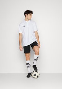 Hvid polo-shirt med en tre-knap knaplukning og sort Adidas-logo, parret med sorte shorts og mønstrede hvide knæstrømper, stående på en fodbold.