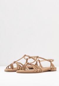 Sandales en suède beige avec des lanières entrecroisées et des accents métalliques. Semelle plate avec une bride de cheville réglable. Design ouvert au niveau des orteils.
