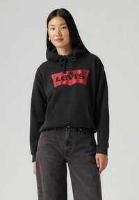 Sudadera negra de tela suave, con un logo rectangular rojo de Levi's en el frente, capucha con cordón y bolsillo tipo canguro. Combinada con jeans grises.