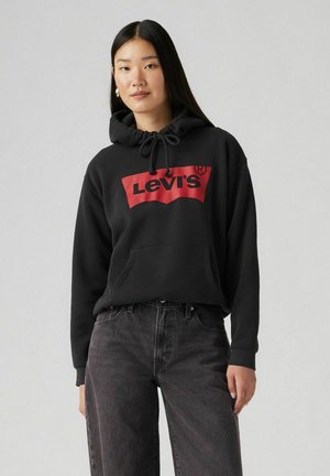 Black hoodie made of soft fabric, featuring a red rectangular Levi's logo on the front, drawstring hood, and kangaroo pocket. Worn with gray jeans.