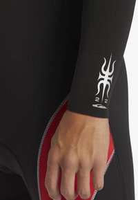 Svart våtdräkt med röda accenter och silverstickningar. Har en logotyp på handleden. Neoprenmaterial för flexibilitet och värme.