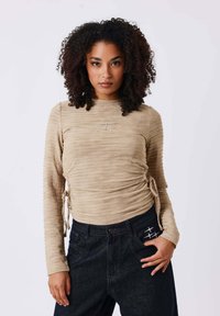 Maglia a maniche lunghe beige in tessuto testurizzato, con dettagli arricciati sui lati e un piccolo logo ricamato sul petto. Abbinata a jeans scuri.