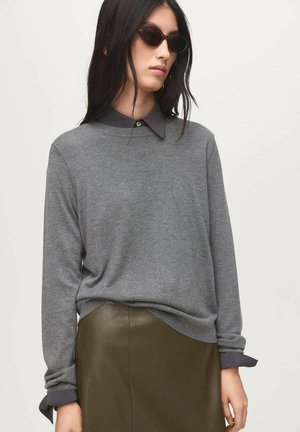 Pull gris avec un col rond, accompagné d'une chemise à col sombre en dessous. Manches longues avec des poignets retroussés, porté avec une jupe vert olive.