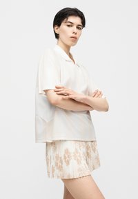 Νεαρό άτομο με κοντά σκούρα μαλλιά που φοράει λευκό ανοιχτό μανίκι κοντό πουκάμισο και πλισέ φούστα με μπεζ λουλουδάτο μοτίβο.