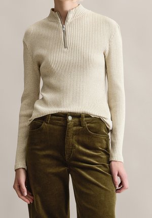 Beiger, gerippter Langarm-Pullover mit kurzem silbernen Reißverschluss, kombiniert mit olivgrünen Cordhosen.