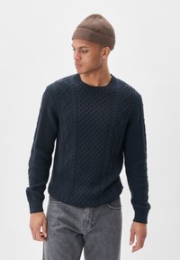 Mornarsko pleteno pulover s teksturiranimi vzorci in rebrastimi manšetami, v kombinaciji s sivo denim hlačami in rjavim pletenim klobukom.