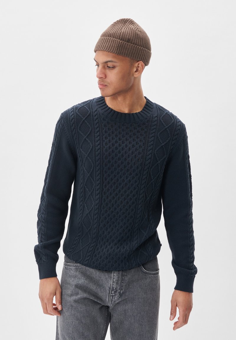 Mornarsko pleteno pulover s teksturiranimi vzorci in rebrastimi manšetami, v kombinaciji s sivo denim hlačami in rjavim pletenim klobukom.