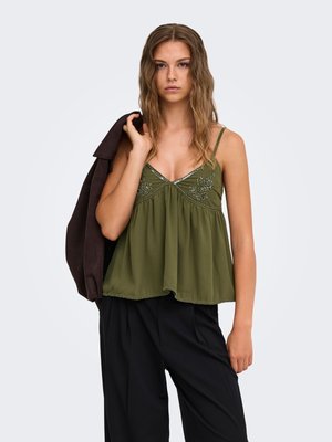 Joven mujer con cabello ondulado lleva una camiseta sin mangas verde oliva y pantalones negros, sosteniendo una chaqueta oscura sobre el hombro contra un fondo liso.