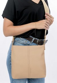 Borsa a tracolla in pelle beige con una superficie liscia, una singola cucitura verticale e una tracolla in canvas regolabile; indossata sopra una maglietta nera e jeans.