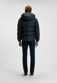 Mörkblå pufferjacka med huva, med ett quiltat mönster och elastiska ärmar, bärs över mörkblå byxor och skor, sett från baksidan.