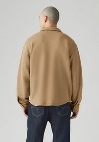 Bruine overshirt met een ontspannen pasvorm, lange mouwen en knoopmanchetten. Heeft een minimalistisch ontwerp en een gladde textuur, gedragen met donkere jeans.