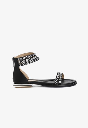 Black sandal featuring metallic stud accents, ankle strap with buckle, flat sole, and a silver rectangular heel. Textured leather design.