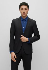 Completo nero con rever a punta e due tasche frontali, abbinato a una camicia blu a pois. Tessuto liscio, design aderente, che mette in risalto le maniche.