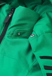 Grüne isolierte Jacke mit einer Reißverschlusstasche, metallischem Schnappverschluss, strukturiertem Stoff und kontrastierenden schwarzen Klettakzenten am Ärmel.