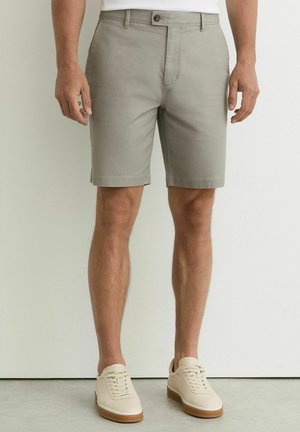 Mees, kes kannab helehallist kohandatud lühikseid pükse ja beeže tossusid, seisab valge seina vastu, käed lõdvestunult külgedel.