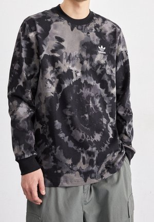 Person iført en langærmet sort og grå tie-dye Adidas-trøje med et lille hvidt logo på brystet og grønne cargobukser.