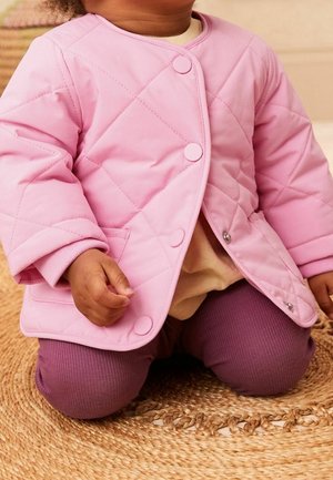 Giacca trapuntata rosa con bottoni rotondi e una texture morbida, indossata su una maglia beige e pantaloni viola a coste, seduta su una superficie intrecciata.