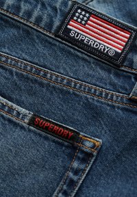 Tissu en denim bleu avec des coutures jaunes. Présente une étiquette rectangulaire Superdry avec un design de drapeau américain et un texte rouge sur fond noir.