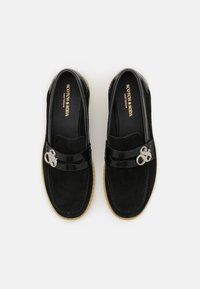 Schwarze WildlederSlipper mit einem PatentschreiberRiemen, der ein silbernes Logo-Detail aufweist. Strukturierte Sohle mit einem kontrastierenden hellen Rand.