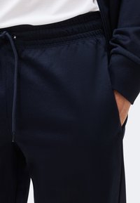 Άτομο που φοράει σκούρο μπλε φόρμα με κορδόνι και σκούρο μπλε μπουφάν με φερμουάρ, με το χέρι στην τσέπη, συνδυασμένο με λευκό πουκάμισο.