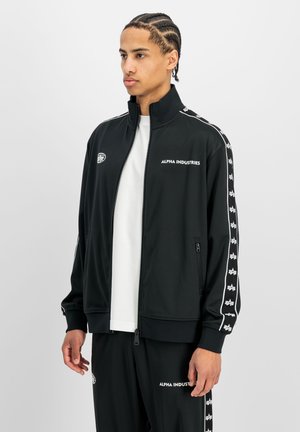 Jonge man met gevlochten haar, gekleed in een zwart Alpha Industries trainingspak met witte logodetails en een wit T-shirt, staande tegen een effen achtergrond.