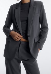 Person in einem dunkelgrauen Anzugjackett und -hose mit schwarzem Oberteil, der die Jacke anpasst und dabei ein kleines Detail des Bauches zeigt.