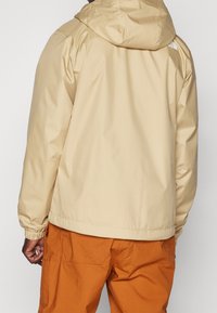 Man wearing beige hooded jacket and orange pants, shown from the back against a plain background.