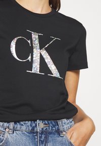 Czarny bawełniany T-shirt z dużym iryzującym logo "CK". Krótkie rękawy i klasyczny krój. Noszony z niebieskimi dżinsami.