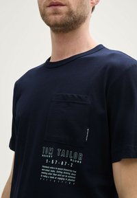 Camiseta de algodón azul marino con mangas cortas, cuello redondo y un bolsillo en el pecho. Presenta un logotipo impreso y texto en blanco en contraste.