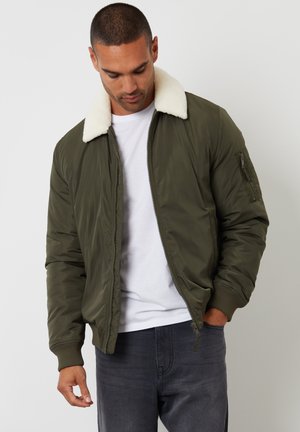 Chaqueta bomber de color verde oliva con un cuello de piel sintética blanca, cierre delantero con cremallera, puños de canalé y un bolsillo con cremallera en la manga izquierda.