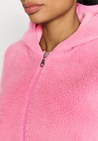 Różowa bluza z kapturem z mikropolaru o teksturowanej, puszystej powierzchni. Posiada zamek błyskawiczny na całej długości oraz kaptur. Suwak zamka ma okrągły uchwyt.