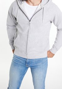 Sweat à capuche zippé gris en tissu doux, avec une capuche à cordon de serrage et des poches avant, associé à un jean en denim bleu clair.